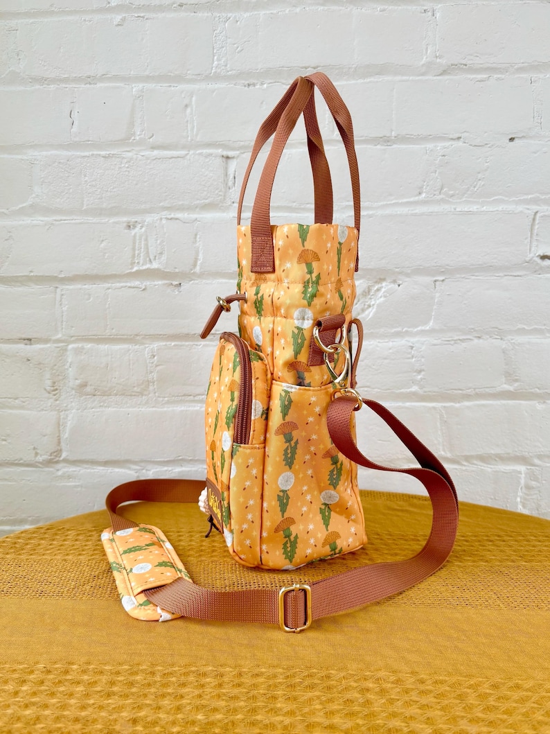 Op de afbeelding: Een kleine, gele en bruine tote bag met een bloemenprint. De tas heeft een bovenste handvat en een lange verstelbare schouderband. De tas heeft een ritsvak aan de voorkant en een klein vak aan de zijkant.