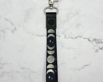 May include: A black key chain with a silver clip and a moon phase design. The chain has a silver metal ring at the top and a black fabric strap with a white moon phase design.