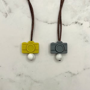 May include: Two camera-shaped silicone charms on brown cords with white marble beads. The camera charms are yellow and gray.