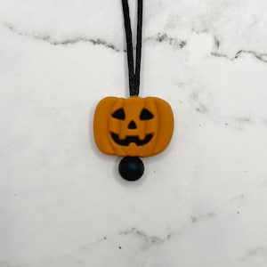 以下が含まれることがあります： 黒いコードネックレスに、オレンジ色のシリコン製かぼちゃのチャームと黒いシリコンビーズが付いている。
