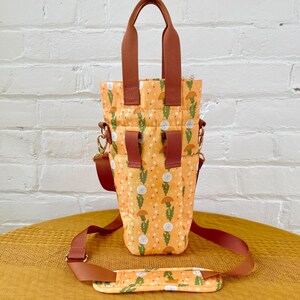 Op de afbeelding: Een bruine en oranje tote bag met een paardenbloemprint. De tas heeft een bovenste handvat en een lange verstelbare schouderband.