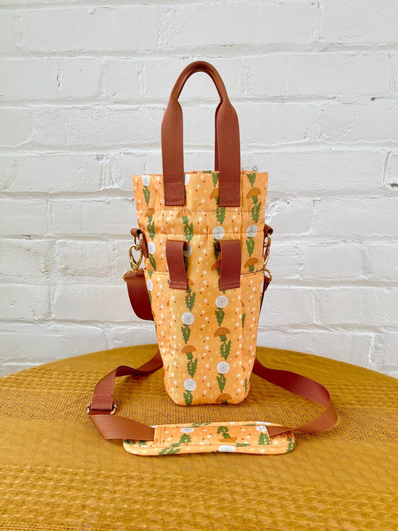 Op de afbeelding: Een bruine en oranje tote bag met een paardenbloemprint. De tas heeft een bovenste handvat en een lange verstelbare schouderband.