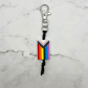 May include: A rainbow pride flag keychain with a silver key ring and black cord. The flag features the traditional rainbow colors with additional brown, black, and white stripes.