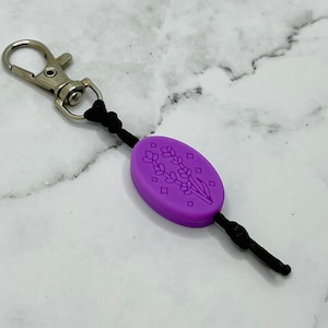 May include: Purple silicone keychain with a lavender flower design and a silver metal clasp.