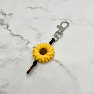 May include: A yellow sunflower charm with a brown center attached to a brown cord and a silver key chain.