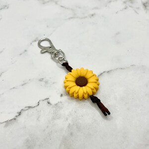 May include: A yellow sunflower charm with a brown center attached to a brown cord with a silver key ring.