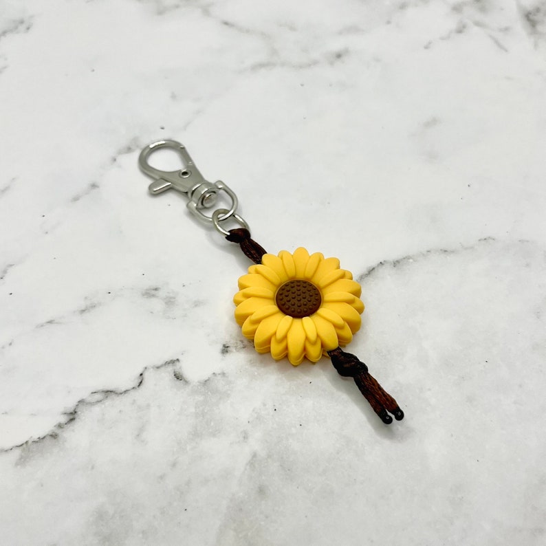 May include: A yellow sunflower charm with a brown center attached to a brown cord with a silver key ring.