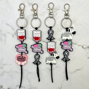 May include: Three black keychains with different charms. The charms include a pink heart with the text "Don't be so hard on yourself", a zebra, and a black and white ribbon. Each charm also includes a pink and red IV bag with the text "Supplement energy" and a pink pill with the text "It's ok if you need meds everyday".