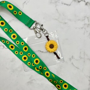 May include: A green lanyard with a sunflower pattern and a clear plastic ID badge holder. A small sunflower charm hangs from the lanyard.