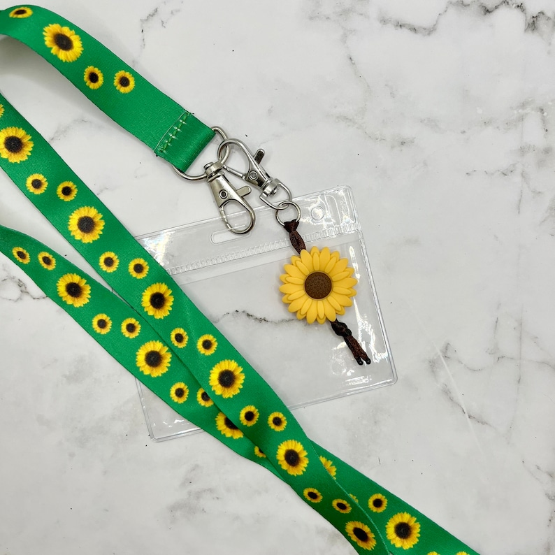 May include: A green lanyard with a sunflower pattern and a clear plastic ID badge holder. A small sunflower charm hangs from the lanyard.
