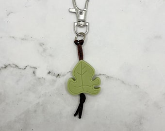 May include: A green leaf-shaped keychain with a brown cord and a silver metal clasp.