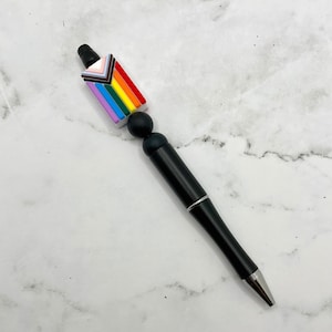 May include: A black pen with a rainbow pride flag charm. The charm is made of colorful beads and features a white background with a rainbow chevron design.