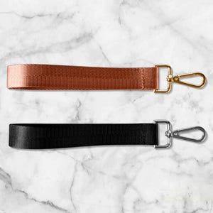Puede incluir: Dos correas de muñeca: una en color cobre-bronce con un cierre dorado, y otra negra con un cierre plateado. Ambas correas están hechas de material tejido y están diseñadas para sujetar llaves o bolsos pequeños.
