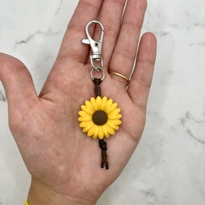 May include: A yellow sunflower charm keychain with a brown center and a brown cord attached to a silver metal clasp.