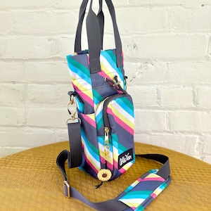 May include: A small, striped, gray, blue, pink, yellow, and white crossbody bag with a top handle. The bag has a zippered pocket and a small sunflower charm. The bag is made by the brand "Artisan".