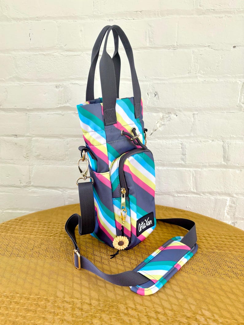 May include: A small, striped, gray, blue, pink, yellow, and white crossbody bag with a top handle. The bag has a zippered pocket and a small sunflower charm. The bag is made by the brand "Artisan".
