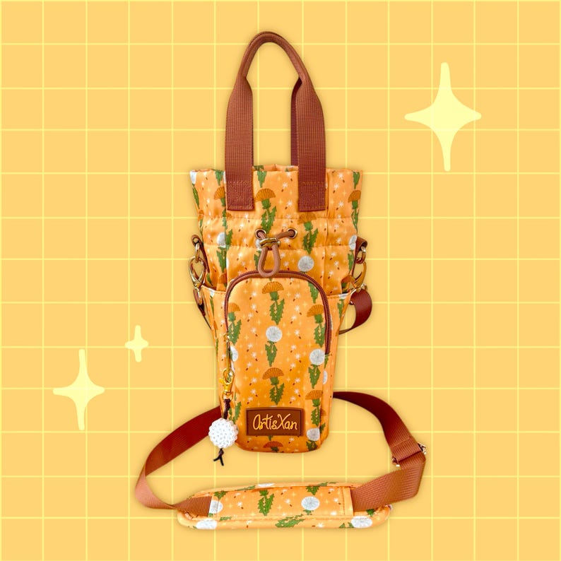 Op de afbeelding: Een bruine en oranje tote bag met een paardenbloemprint en de tekst "Arti&Yan". De tas heeft een bruine band en een ritssluiting.