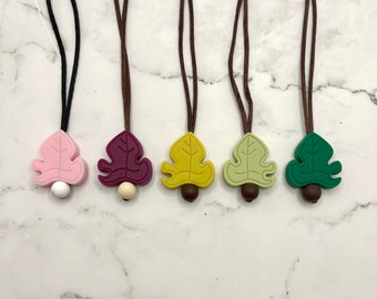 May include: Five leaf-shaped silicone necklaces with brown cords. The leaves are in various colors: pink, burgundy, yellow, green, and dark green. Each leaf has a small brown bead at the bottom.