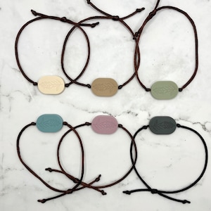May include: Six brown cord bracelets with oval-shaped charms featuring a floral design. The charms are in various colors, including beige, light brown, green, blue, and purple.