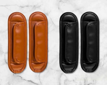 Op de afbeelding: Twee paar leren schouderpads. Eén paar is warm bruin, de andere is zwart. Elke pad heeft een gebogen ontwerp met een gecentreerd gewatteerd gedeelte en een lus voor bevestiging, voor comfort en ondersteuning.