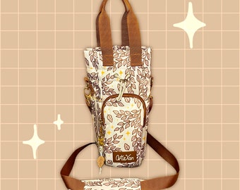 May include: A brown and white canvas tote bag with a leafy pattern and gold accents. The bag has a top handle and a long adjustable strap. The bag has a small pocket on the front with the text "Atle Xan" printed on it.