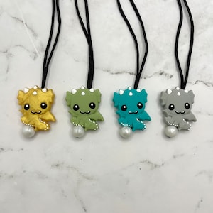 May include: Four necklaces with different colored dragon charms. The charms are made of silicone and have a pearl bead at the bottom of the necklace. The necklaces are black.