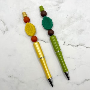 May include: Two pens with fall-themed decorations. One pen has brown, orange, and yellow beads with a yellow leaf charm. The other pen has brown, green, and brown beads with a green leaf charm.