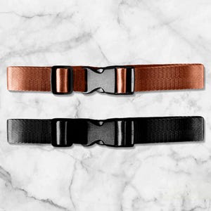 Puede incluir: Dos cinturones ajustables de cincha de nailon con hebillas de plástico. Uno es de color cobre y el otro negro. Los cinturones están colocados sobre un fondo de mármol blanco y gris.