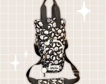 May include: A black and white tote bag with a crescent moon pattern. The bag has a top handle and a long adjustable shoulder strap. The bag has a small zippered pocket on the front. The bag has a white tag with the text "ArtisXan" on it.