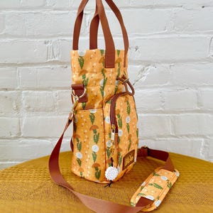 Op de afbeelding: Een kleine, gele en groene geprinte tote bag met een bruine band en een witte pompon-bedel. De tas heeft een ritsvak aan de zijkant en een bovenste handvat.