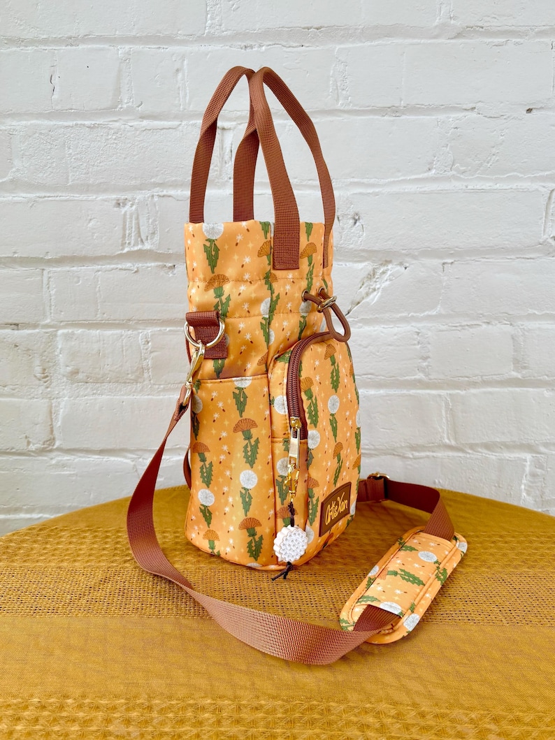 Op de afbeelding: Een kleine, gele en groene geprinte tote bag met een bruine band en een witte pompon-bedel. De tas heeft een ritsvak aan de zijkant en een bovenste handvat.