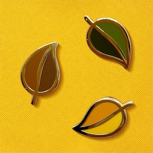 May include: Three leaf-shaped enamel pins with gold-colored borders. One pin is brown, another is green, and the third is yellow and orange. The pins are arranged on a yellow background.