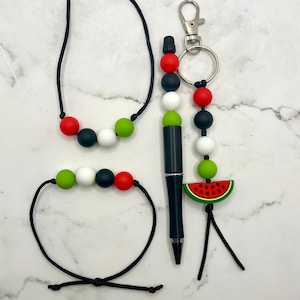 Puede incluir: Un conjunto de tres collares de cuerda negra con cuentas de silicona de colores. Los collares presentan cuentas rojas, verdes, blancas y negras. Un collar tiene un dije de sandía y un llavero.