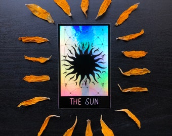 Puede incluir: Una carta de tarot con una ilustración de sol negro sobre un fondo holográfico. La carta está rodeada de pétalos de flores secas sobre una superficie negra. El texto en la carta dice "THE SUN".