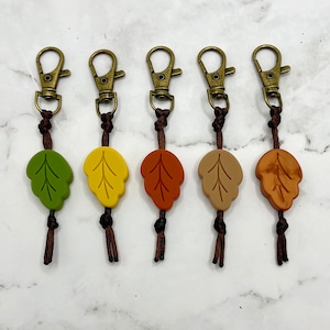 May include: Five keychains with different colored leaf charms. The charms are green, yellow, orange, brown, and red. Each keychain has a gold-colored metal ring and a brown cord.