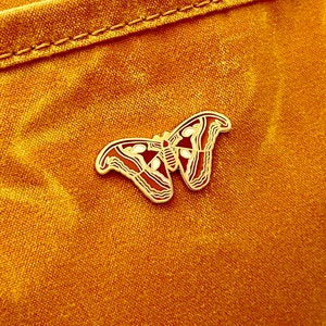 May include: A gold-coloured butterfly pin with red and white accents. The pin is attached to a mustard yellow fabric background.
