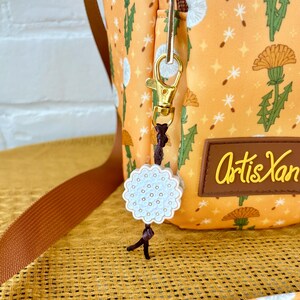 Op de afbeelding: Een kleine oranje en witte stoffen tas met een paardenbloemprint. De tas heeft een gouden rits en een gouden clip met een witte keramische paardenbloembedel eraan. De tas ligt op een bruin en wit geweven oppervlak. De tekst "Artis Yan" staat op de tas gedrukt.