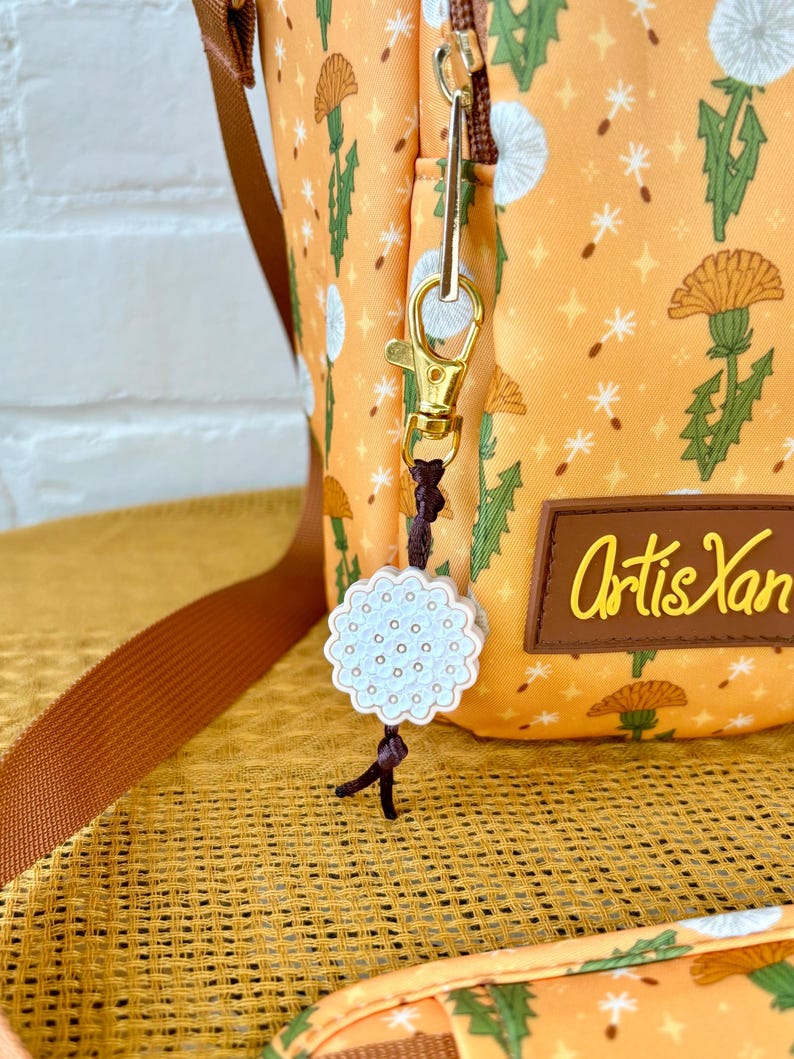 Op de afbeelding: Een kleine oranje en witte stoffen tas met een paardenbloemprint. De tas heeft een gouden rits en een gouden clip met een witte keramische paardenbloembedel eraan. De tas ligt op een bruin en wit geweven oppervlak. De tekst "Artis Yan" staat op de tas gedrukt.