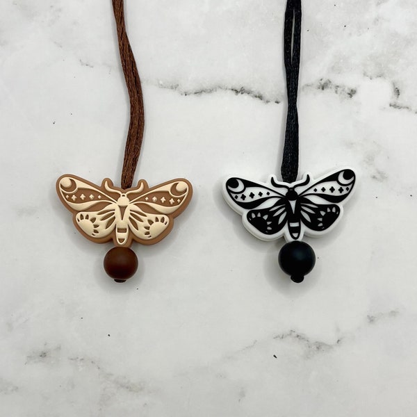 May include: Two necklaces with a brown and black cord and a silicone pendant shaped like a moth with intricate details. The moth pendant is white with black details on the brown cord and black with white details on the black cord.