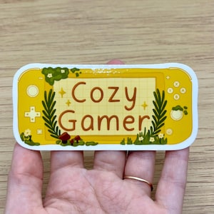 May include: Yellow and white sticker with a cartoon illustration of a Nintendo Switch console. The console has a grid pattern on the screen and the text "Cozy Gamer" on the screen.