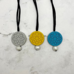 Puede incluir: Tres collares con colgantes que presentan un diseño circular de un sol y fases lunares. Los colgantes son grises, amarillos y azules.