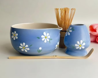 May include: A blue ceramic matcha tea set with white daisy floral designs. The set includes a bowl, whisk, whisk holder, and scoop. The bowl has a spout for easy pouring. The whisk is made of bamboo.