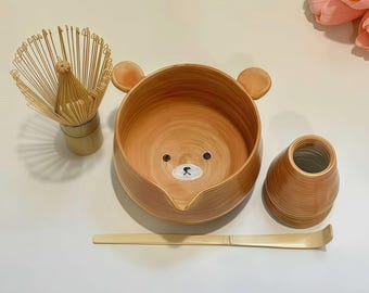 Può includere: Un set da tè matcha in ceramica marrone chiaro con una ciotola a forma di orso con orecchie e un viso dipinto. Il set include una frusta di bambù, un contenitore per il tè e un cucchiaio di bambù. La ciotola misura circa 15 cm di diametro.