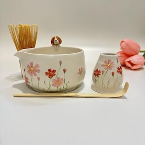 Puede incluir: Un juego de té matcha de cerámica color crema con un cuenco y un pequeño jarrón a juego, ambos adornados con diseños florales rosas y rojos. Una pequeña figura de perro se encuentra en el borde del cuenco. Incluye un batidor y una cuchara de bambú.