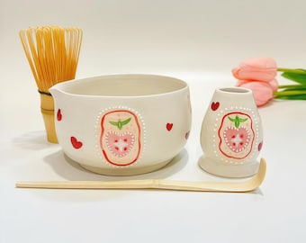 May include: A ceramic matcha bowl and a whisk holder, both off-white with hand-painted pink strawberry motifs and small red hearts. A bamboo whisk and a bamboo tea scoop are included. The strawberry designs feature white dotted borders.