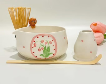May include: A ceramic matcha bowl with a hand-painted pink oval frame containing green leaves and pink flowers, featuring a small brown dog figurine on the rim, accompanied by a bamboo whisk, a bamboo scoop, and a white ceramic whisk holder.