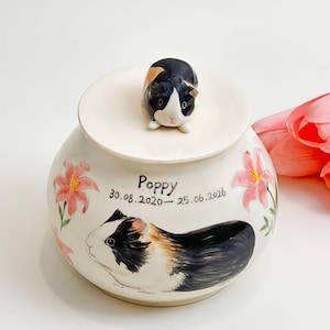 Op de afbeelding: Een witte keramische pot met een deksel voorzien van een handgeschilderd, driekleurig caviafiguur. De pot toont een geschilderde driekleurige cavia, roze lelies en de tekst "Poppy 30.08.2020 - 25.06.2026" in zwarte letters.