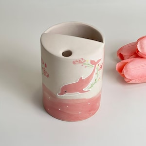 May include: A cylindrical ceramic vase with a light pink base and a dolphin illustration. The dolphin is pink and surrounded by floral accents. The vase has a curved top and a small hole. Pink tulips are in the background.