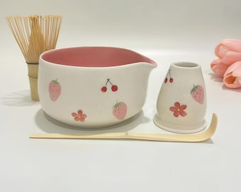 May include: A matcha tea set featuring a white ceramic bowl with a pink interior and strawberry and cherry designs. Includes a bamboo whisk, a small ceramic holder, and a bamboo scoop. The set is designed for preparing matcha tea.