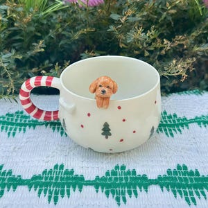 Op de afbeelding: Een witte keramische mok met een rood-wit gestreepte handgreep. De mok heeft een klein bruin hondje op de rand, met rode stippen en groene kerstboomontwerpen. De mok staat op een wit en groen patroon.